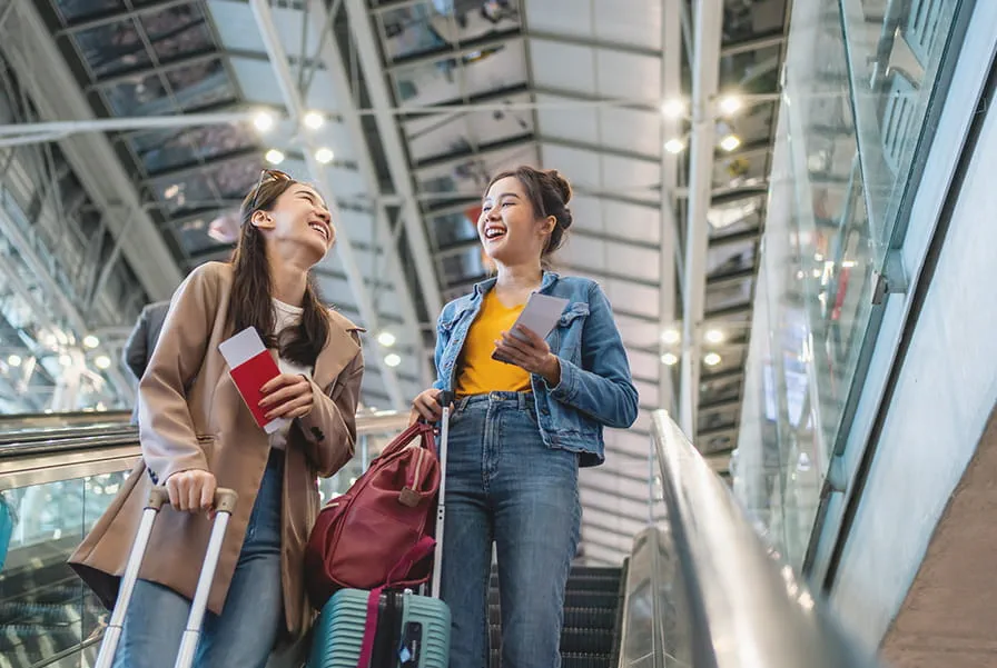 Two smiling women on an airport escalator with luggage and travel passes.
