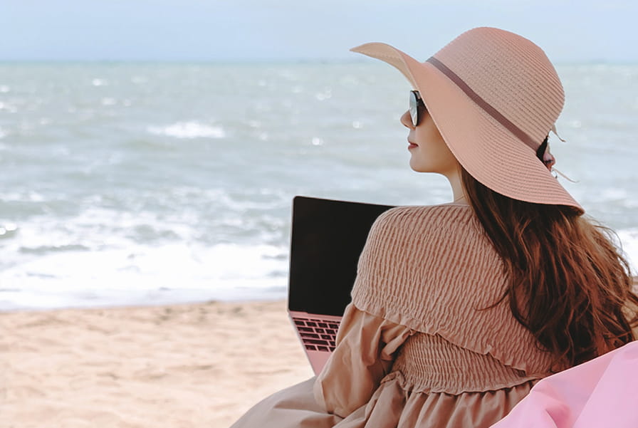 Woman wearing a wide-brimmed hat and sunglasses, sitting on a beach chair with a laptop while looking out at the ocean.
