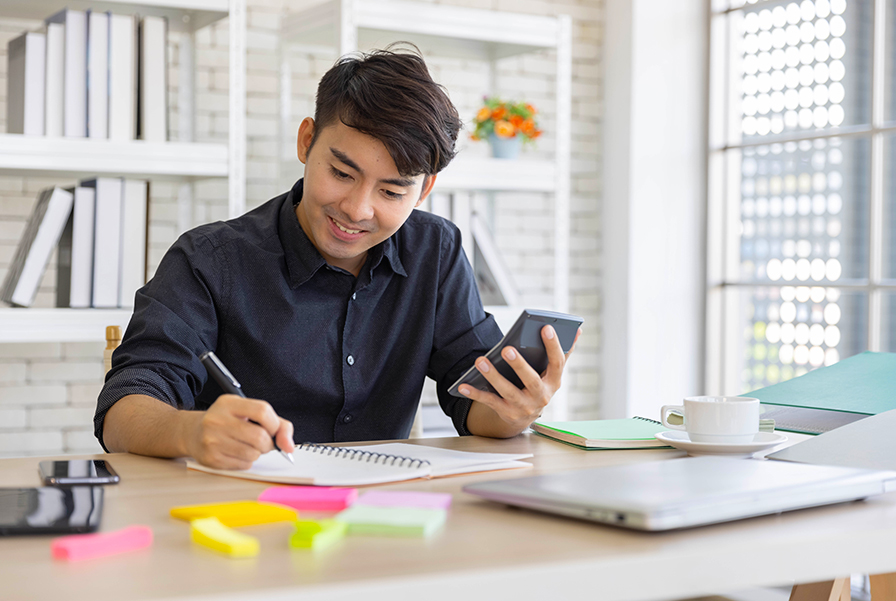 Man working at a desk while checking his phone and writing notes, representing everyday money management using a account.
